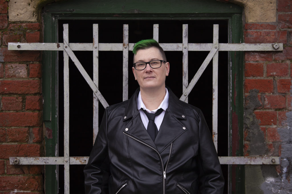 AJ Fitzwater, a white person with green hair, short on top, shaved on sides. They are wearing black rimmed glasses, a black leather jacket, white shirt, and black tie. They stand with their hands in pockets, in front of silver cross bars of a window attached to red brick, as they look straight ahead.